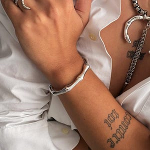 Op de afbeelding: Een zilverkleurige sieradenset bestaande uit een dikke ketting met een gebogen hanger in bamboestijl, een bijpassende armband, een ring met meerdere banden en een slangvormige ring, gedragen bij een wit overhemd met knoopsluiting.