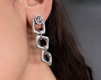 May include: A silver-toned drop earring featuring three connected square-shaped links of varying sizes, with the top link containing a small circular accent. The earring hangs vertically from the earlobe, showcasing a polished metallic finish.