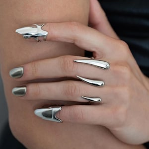 Könnte beinhalten: Silberfarbene Ringe und ein Armband. Die Ringe haben ein einzigartiges klauenartiges Design, wobei einige länglichen Tropfen ähneln. Das Armband hat einen großen, kreisförmigen Verschluss und ein Kettendesign. Die Nägel sind silberfarben lackiert.