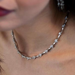 May include: A silver-toned metal necklace featuring a series of evenly spaced, small, clear, faceted stones set along the chain. The necklace rests against the neck, showcasing a repeating pattern of metallic links and embedded sparkling stones.