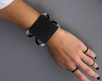 Op de afbeelding: Een zwarte vierkante armband met een centrale matzwarte plaat verbonden door zwarte elastische banden en zilverkleurige metalen kralen. De armband wordt om de pols gedragen, vergezeld van drie dunne zwarte ringen aan de vingers van de hand.