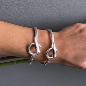 May include: Two silver-toned metal bracelets featuring a toggle-style clasp mechanism and two matching silver-toned rings with a V-shaped geometric design. The jewelry pieces have a smooth, polished finish and a consistent metallic appearance.