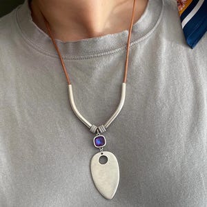 May include: A necklace featuring a brown leather cord, two curved silver-toned metal tubes, a small square purple crystal, and a large, smooth, teardrop-shaped silver-toned pendant with a circular cutout near the top center of the piece.