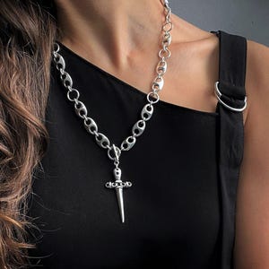 May include: A silver-toned necklace featuring a thick chain with oval links and a central dagger-shaped pendant. The necklace is paired with a matching silver-toned bracelet composed of small round beads and a larger cylindrical center piece.