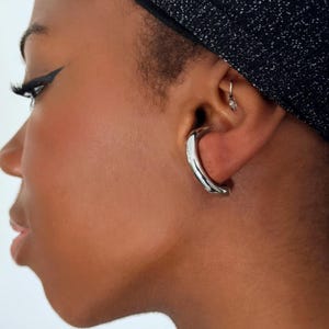 Puede incluir: Un pendiente plateado y curvado se lleva en la oreja. El pendiente tiene una superficie lisa y pulida y un diseño minimalista. La persona lleva una diadema negra con una textura brillante.