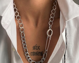 May include: A silver-toned chain necklace featuring a mix of hexagonal and oval links, integrated with a large safety pin clasp mechanism. The necklace is worn over a crisp white collared button-down shirt, showcasing a metallic finish and industrial design.