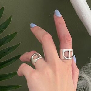 Könnte beinhalten: Zwei silberne Ringe mit ausgeschnittenem Ankerketten-Design an Zeige- und Ringfinger, kombiniert mit einem silbernen Perlenarmband und einem schwarzen Lederband-Armband mit silbernem Metallverschluss und einem silbernen Nietendetail.