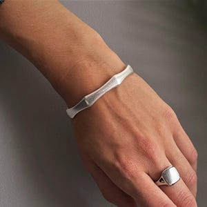 Könnte beinhalten: Ein silberfarbenes Armband mit segmentiertem Bambus-Design und ein passender silberfarbener Siegelring mit einer glatten, polierten quadratischen Oberfläche. Beide Schmuckstücke haben ein mattes, gebürstetes Metall-Finish am Handgelenk.