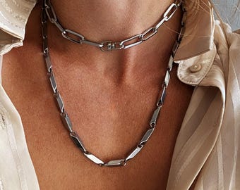 May include: A layered silver-toned necklace set consisting of two distinct chains. The shorter chain features elongated rectangular links, while the longer chain is composed of smaller, segmented metallic links with a polished finish.