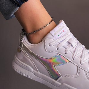 Puede incluir: Una zapatilla blanca con un panel lateral holográfico iridiscente y cierre de cordones blanco a juego. La lengüeta muestra el nombre de la marca Diadora con pequeños detalles circulares en verde, blanco y rojo. Se lleva una tobillera de cadena plateada.