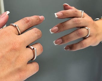 May include: A silver-toned open-ended ring featuring a smooth, cylindrical bar design. The ring is worn on the finger, showcasing a minimalist, polished metallic finish that reflects light, creating a clean and simple aesthetic for everyday wear.