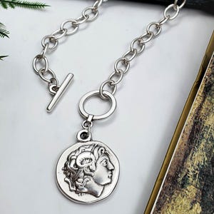 May include: A silver-toned necklace featuring a toggle clasp and a circular pendant. The pendant displays a relief profile of a figure with curly hair. The chain consists of large, rounded links, and the entire piece has a metallic finish.