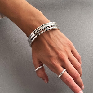 May include: A set of three silver-toned bangles with a smooth, undulating surface texture worn on the wrist, accompanied by two simple, polished silver-toned rings worn on the index and ring fingers, all featuring a reflective metallic finish.