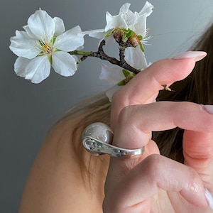 Op de afbeelding: Een zilverkleurige ring met een grote, gladde, bolvormige grijze kraal in een gepolijste metalen band. De ring wordt getoond naast een delicate tak met witte bloesems, wat het contrast tussen de metaalglans en de organische florale elementen benadrukt.