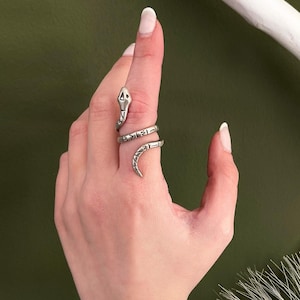 Può includere: Un anello a forma di serpente color argento si avvolge attorno a un dito, caratterizzato da una testa dettagliata e un corpo testurizzato. Un ampio bracciale rigido color argento lucido con superficie martellata riflette la luce.