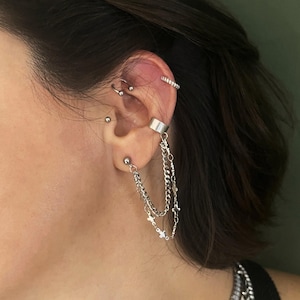 Pode incluir: Um ear cuff prateado conectado a um brinco de pino por duas correntes delicadas. Uma corrente é de elos simples, enquanto a outra apresenta pequenos pingentes em forma de cruz. O ear cuff está posicionado na cartilagem da orelha.