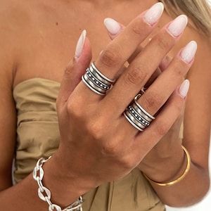 May include: Two wide silver-toned rings featuring a central band with a dotted pattern, worn on the index and ring fingers. A chunky silver-toned chain link bracelet with a toggle clasp is worn on the wrist, alongside a thin gold-toned bangle.
