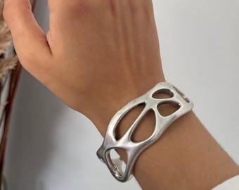 以下が含まれることがあります： オープンで有機的なデザインのシルバーカラーのカフブレスレットが手首に着用されています。テクスチャード加工されたバンドが付いたお揃いのシルバーリングが指にあります。背景は、乾燥した植物のあるニュートラルな白です。