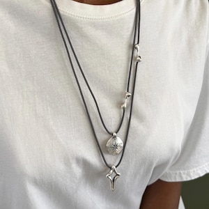 Peut inclure: Un collier à plusieurs rangs avec un cordon gris foncé. Le collier comporte trois pendentifs argentés : une rose des vents, une étoile filante et de petites perles décoratives. Le collier est porté sur un t-shirt blanc.