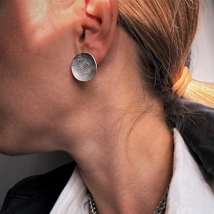May include: A silver-toned circular stud earring with a textured surface and a silver-toned chain necklace featuring a toggle clasp closure and a small, round, blue crystal pendant hanging from the centre of the toggle mechanism.
