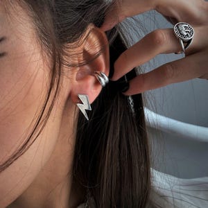 Op de afbeelding: Een zilverkleurige bliksemschicht-oorsteker, een gladde zilveren oormanchet en een zilveren ring met een rond vlak voorzien van een gedetailleerd rasterpatroon en een centrale letter E, allemaal gedragen aan het oor en de vinger.