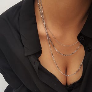 May include: A layered silver-toned necklace consisting of three distinct chains of varying lengths and link styles. The chains are worn together, featuring a smooth snake chain and two textured link chains, resting against a black collared shirt.