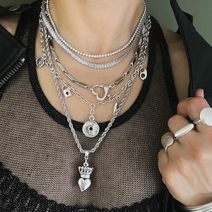 May include: A layered silver-tone necklace set featuring a crystal tennis chain, a chain with a heart-shaped clasp, a padlock charm, a circular coin pendant, and a heart pendant topped with a crown. Three silver rings and a stack of silver bangles.
