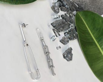 May include: A pair of silver-toned earrings featuring two distinct designs. One earring is shaped like a large safety pin with a smooth finish, while the other consists of a vertical chain of small, interconnected circular links with a polished surface.