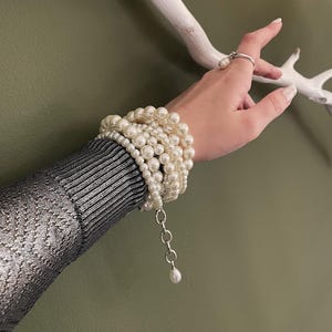 May include: A multi-strand bracelet composed of cream-colored spherical pearls of varying sizes, featuring a silver-toned metal chain extender with a single dangling pearl charm, and a matching silver-toned ring with a small pearl accent.