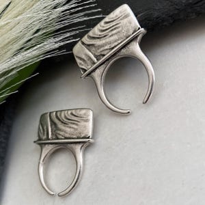 May include: Two silver-toned rings featuring a rectangular top section with a raised, swirling, wood-grain-like texture. Each ring has an open, adjustable band design with a smooth, polished finish on the lower portion of the ring structure.