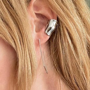 May include: A woman with blonde hair wearing a silver ear cuff and a dangling silver earring. The ear cuff is a thick, textured piece that wraps around the ear. The earring is a thin, straight line. The woman is also wearing a silver necklace and a denim jacket.