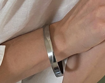 Könnte beinhalten: Ein weißes Hemd mit Knopfleiste, kombiniert mit Silberschmuck, bestehend aus einer mehrlagigen Kugelkette, einem glatten, geschwungenen Ring, einem Ring mit strukturierter Gliederkette und einem polierten Silberarmreif am Handgelenk.