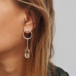 May include: A silver-toned earring featuring a circular hoop attached to a vertical bar, which connects to a rectangular pendant with a cutout cross design. The earring is worn in the earlobe, while a small black hoop is visible in the upper ear.