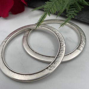 May include: Two circular silver-toned bangles featuring a textured, embossed pattern along the outer surface. The bracelets have a smooth inner band and a matte, metallic finish, presented as a pair of matching jewelry pieces with a consistent design.