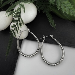 May include: A pair of silver-toned hoop earrings with a teardrop shape. The lower portion of each hoop features a textured, diamond-cut pattern that reflects light, while the upper portion remains smooth and polished with a standard post closure.