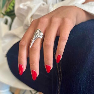 Pu&ograve; includere: Anello color argento con un design ornato e inciso. L'anello ha una forma angolare e appuntita. La mano indossa lo smalto rosso. Un gioiello importante, probabilmente in metallo.