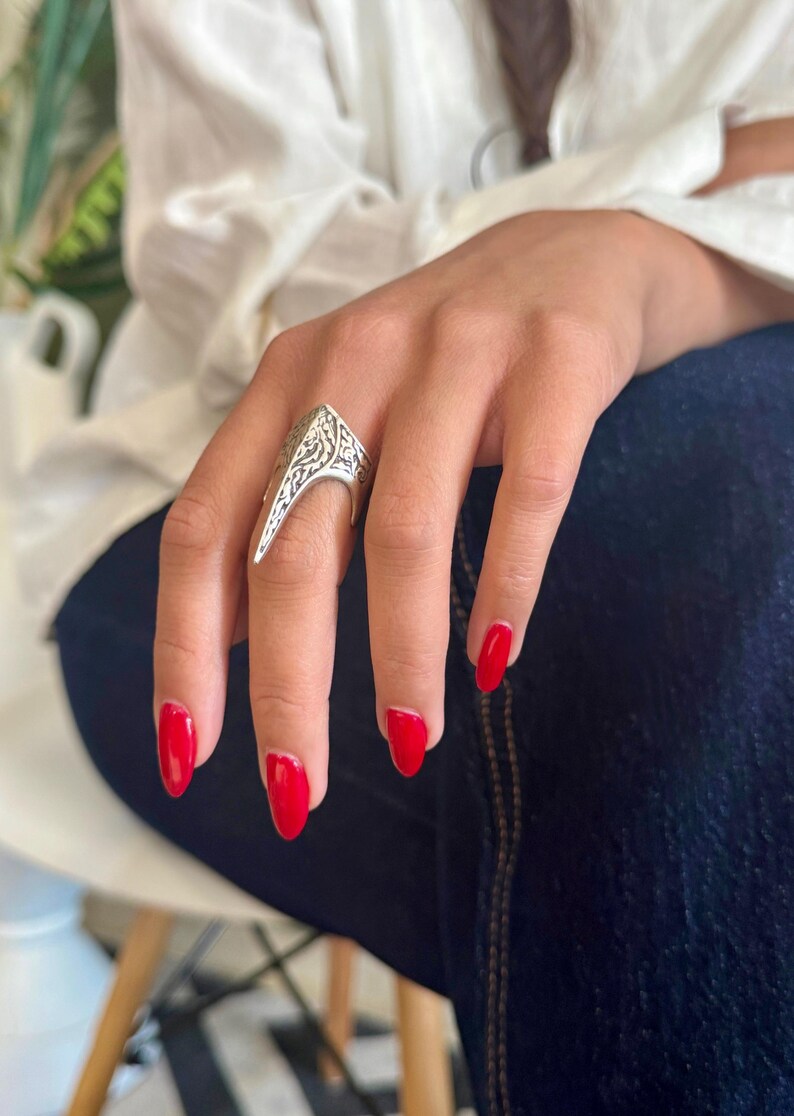 Pu&ograve; includere: Anello color argento con un design ornato e inciso. L'anello ha una forma angolare e appuntita. La mano indossa lo smalto rosso. Un gioiello importante, probabilmente in metallo.