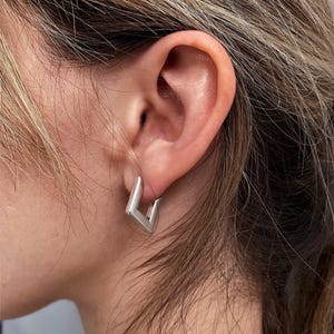 Könnte beinhalten: Ein kleiner, silberfarbener Creolen-Ohrring mit einem markanten, V-förmigen oder kantigen geometrischen Design. Der Ohrring hat eine glatte, metallische Oberfläche und ist am Ohrläppchen befestigt, was eine klare, scharfe Silhouette zeigt.