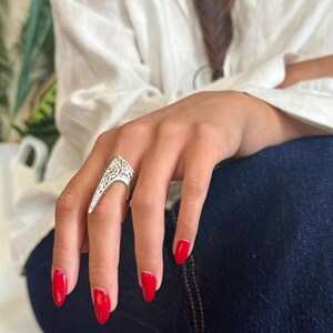 Pu&ograve; includere: Un anello d'argento con un design ornato e un'estensione appuntita. L'anello &egrave; indossato su una mano con smalto rosso. La persona indossa una camicia bianca e jeans blu.