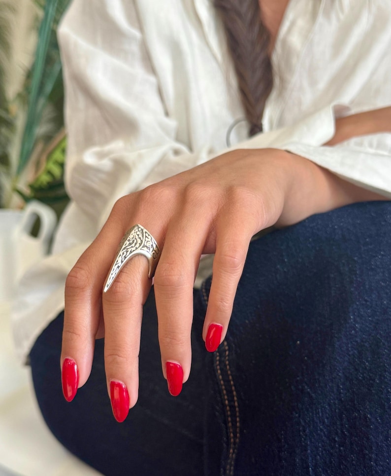 Pu&ograve; includere: Un anello d'argento con un design ornato e un'estensione appuntita. L'anello &egrave; indossato su una mano con smalto rosso. La persona indossa una camicia bianca e jeans blu.