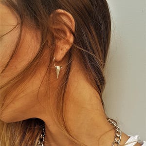 May include: A silver-toned hoop earring featuring a small, textured, cone-shaped pendant hanging from the bottom. The earring is worn in the earlobe, and a thick silver-toned chain necklace is visible around the neck area of the white garment.