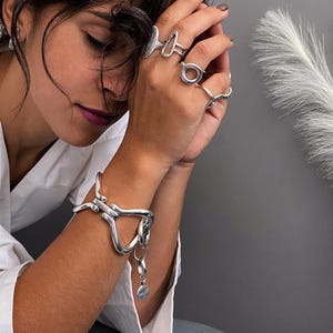 Könnte beinhalten: Ein silberfarbenes Metallarmband mit einem großen, unregelmäßigen, offenen Glieddesign und einem hängenden runden Anhänger, kombiniert mit vier silberfarbenen Ringen an den Fingern, die jeweils verschiedene geometrische Formen wie Kreise und Dreiecke aufweisen.