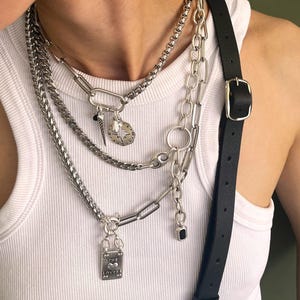May include: A layered silver necklace set with various chain styles and charms. The charms include a compass, a dagger, and a rectangular pendant with the words "The Lovers" and a heart. A black leather strap with a silver buckle is also visible.