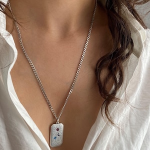 Op de afbeelding: Een zilverkleurige rechthoekige hanger aan een gourmetketting. De hanger is voorzien van een gegraveerd hemels ontwerp met een kleine paarse ronde steen, een donkerblauwe halve maan en een blauwe vierpuntige ster boven een berglandschap.