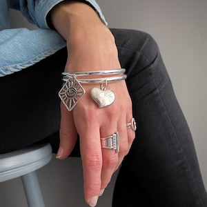 May include: A silver bangle bracelet featuring two dangling charms: one shaped like a diamond with an eye motif and the other a smooth, puffy heart. The bracelet is worn alongside two silver rings, one with a vertical bar design and one simple band.
