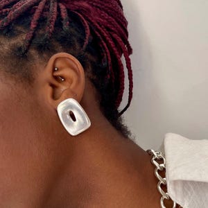 May include: A silver-toned earring with a smooth, rectangular, slightly concave shape featuring a central cutout. The earring has a matte finish and is worn on the earlobe. A thick, silver-toned chain necklace is visible around the neck area.