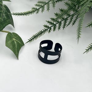 May include: A matte black ring featuring a double band design connected by a vertical bar. The ring has an open back construction, allowing for adjustable sizing. The surface of the metal is smooth and uniform in colour, creating a bold appearance.