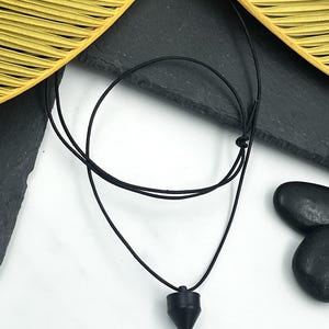Könnte beinhalten: Eine schwarze Anhängerkette mit einem glatten, mattschwarzen konischen Anhänger, der an einer dünnen, verstellbaren schwarzen Kordel hängt. Die Kordel ist mit einem einfachen Knoten versehen, der eine Längenanpassung ermöglicht. Der Anhänger hat eine spitze, geometrische Form.