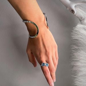 May include: A polished silver-tone open cuff bracelet with a smooth, curved design and a matching silver-tone ring featuring two small, round, blue-toned stones set side-by-side on the band, worn together as a coordinated jewelry set.
