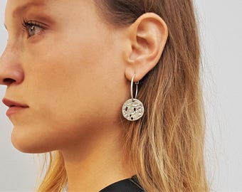 May include: A silver hoop earring featuring a circular pendant with a textured, uneven surface. The pendant has a metallic finish and small, irregular indentations across its face, suspended from a simple, thin silver hoop that passes through the earlobe.
